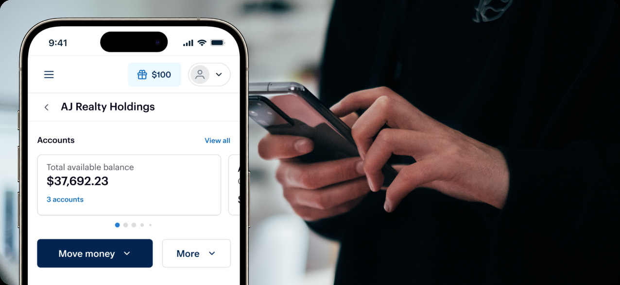 A person using a smartphone is visible in the background. In the foreground, a mobile banking app on another phone screen shows an available balance of $37,692.23 for 'AJ Realty Holdings.' The app includes options to move money and view more accounts.