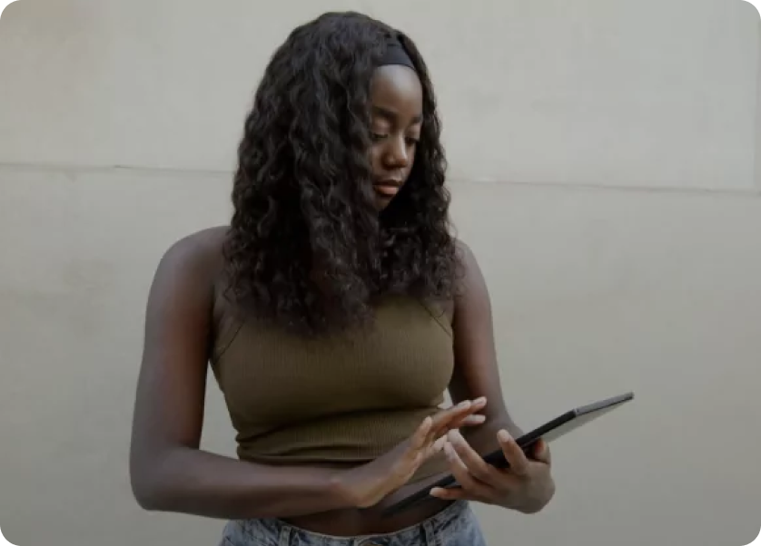 A person with long curly hair, wearing a sleeveless top and jeans, is standing against a plain wall while looking at and tapping on a tablet.