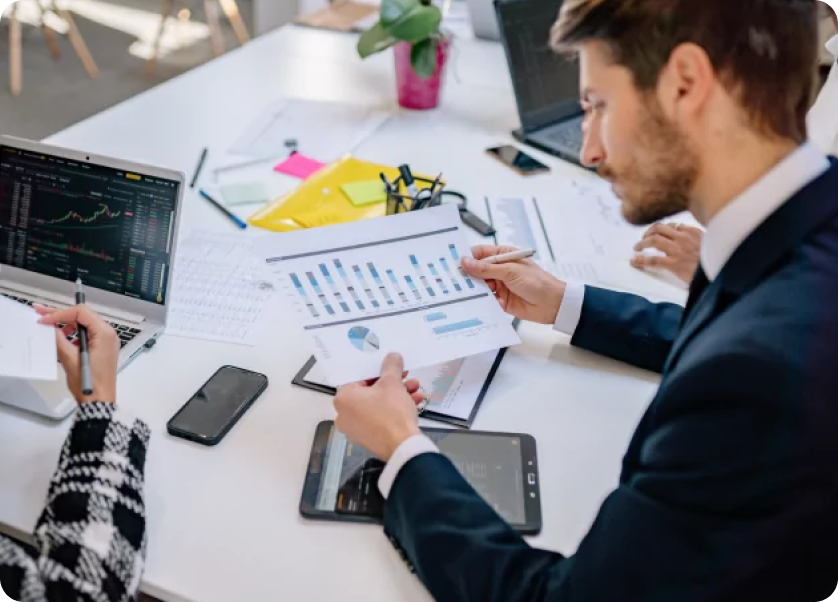 A group of people in business attire are seated at a white table. One person is holding a document with graphs and charts, while another works on a laptop displaying financial data. Office supplies and a plant are on the table.