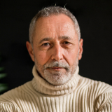 A middle-aged man with short gray hair and a neatly trimmed beard is wearing a light beige turtleneck sweater. He has a serious expression and is set against a dark background with a hint of green foliage on the left side of the image.
