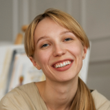A person with blonde hair is smiling warmly at the camera. They are wearing a beige top and are in an indoor setting with a blurred background. The overall atmosphere appears friendly and inviting.