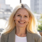 A woman with long blonde hair smiles while facing the camera. She is wearing a checked blazer over a white top. The background shows an urban environment with blurred buildings and greenery. The lighting suggests it is a sunny day.