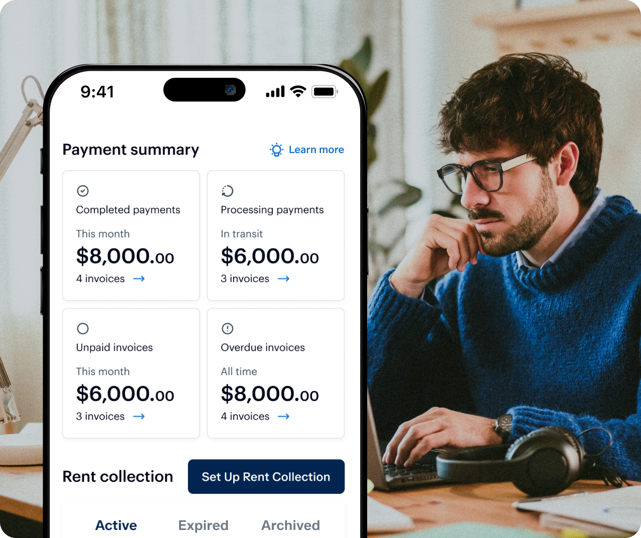 A man with glasses and a beard, wearing a blue sweater, sits at a desk with a laptop. A smartphone screen in the foreground displays a payment summary with details on completed, processing, unpaid, and overdue invoices, featuring amounts and invoice counts.