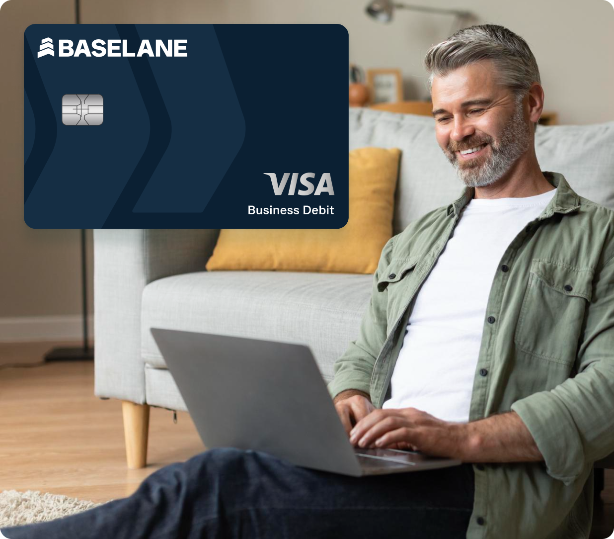 A man with short gray hair and a beard is sitting on the floor, smiling while using a laptop. Above him is an enlarged image of a Baselane Visa Business Debit card. The room has a modern, cozy interior with a sofa in the background.