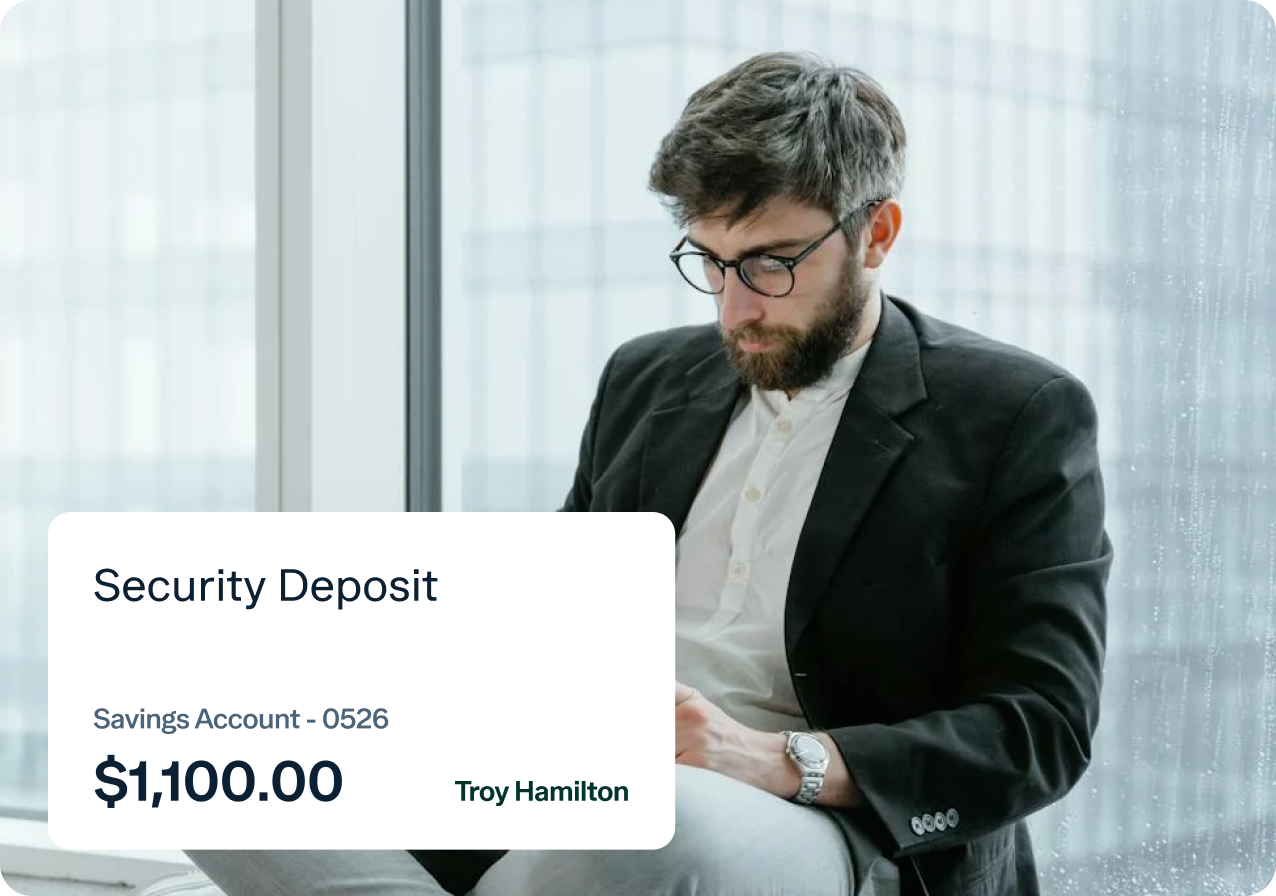 A man with glasses and a beard, dressed in a suit, sits by a window in a high-rise building, focused on his phone. In the foreground, a digital overlay displays "Security Deposit," "Savings Account - 0526," "$1,100.00," and "Troy Hamilton.