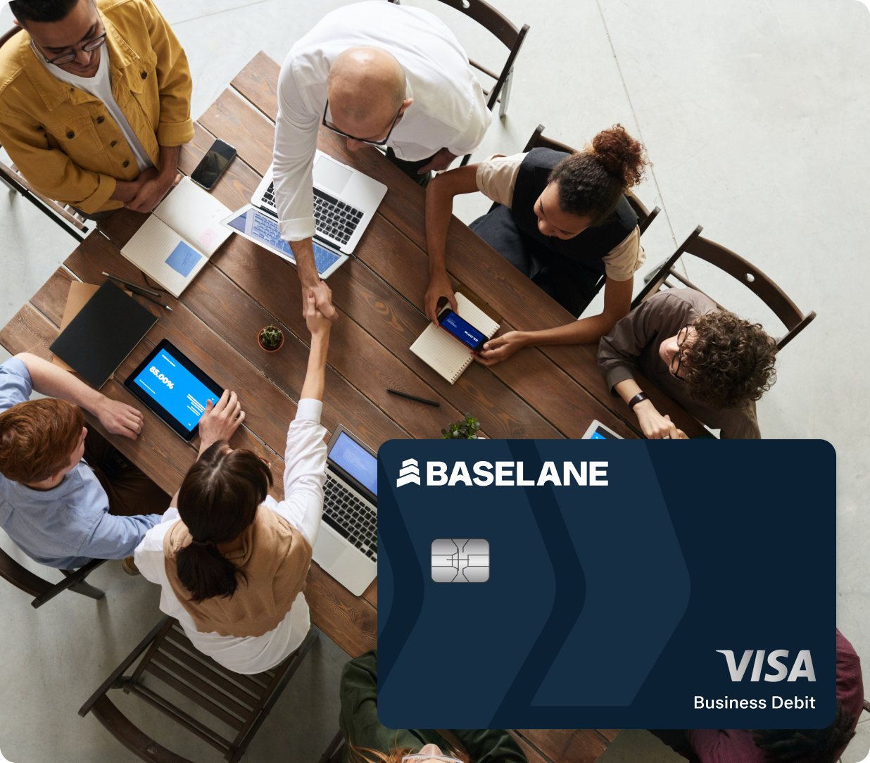 A group of people are gathered around a wooden table, some using laptops and tablets. Two individuals are shaking hands. In the foreground, there is a large, partially transparent Basellane Visa Business Debit card overlaying the image.