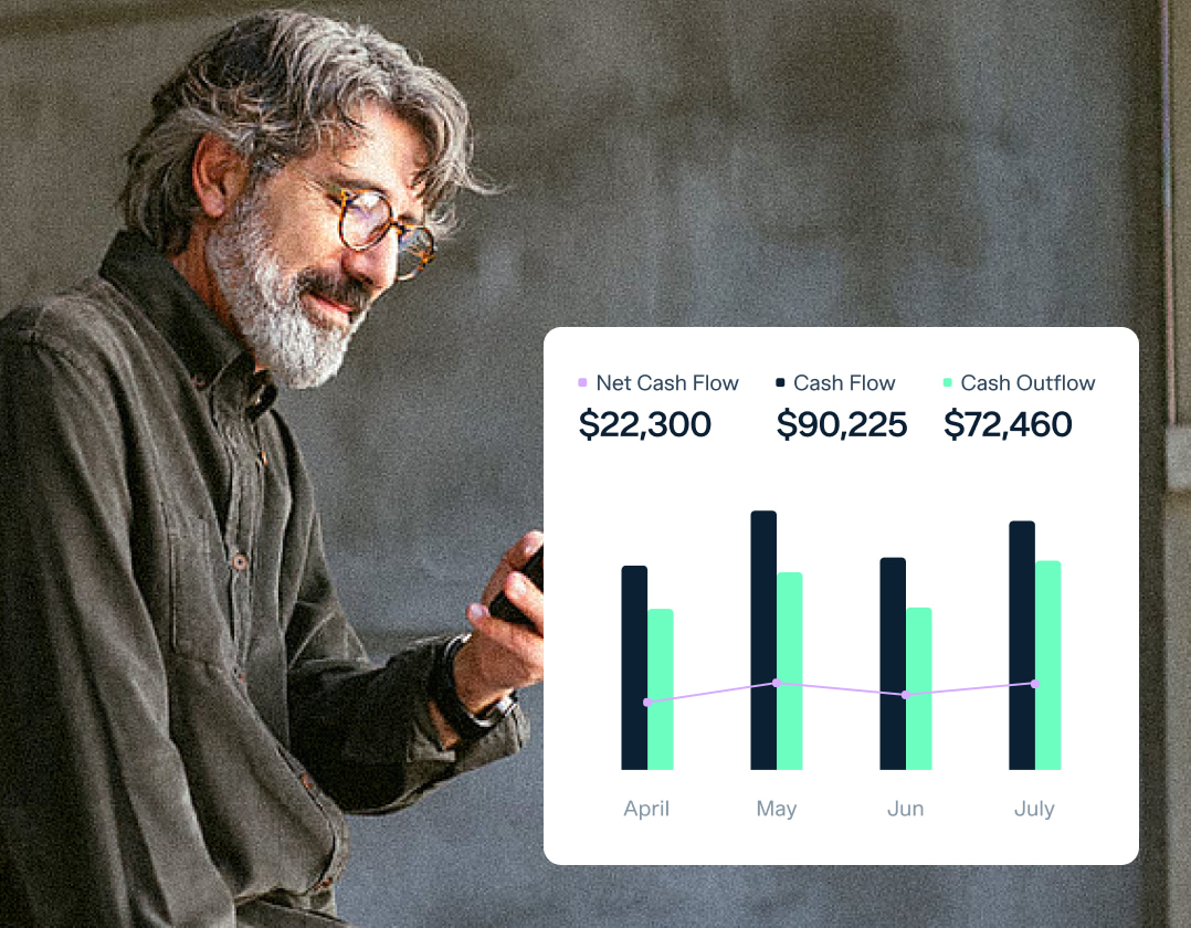 An older man with gray hair and glasses looks at his smartphone while standing beside a concrete wall. An infographic next to him shows a bar and line graph depicting cash flow, net cash flow, and cash outflow for April, May, June, and July.