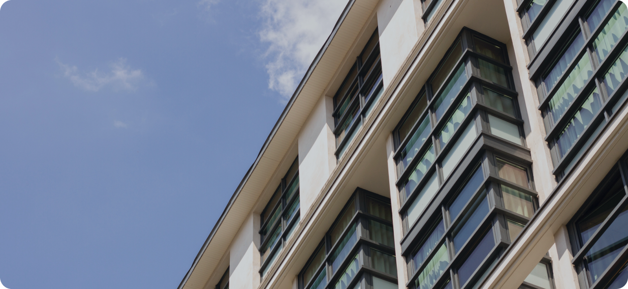 A low-angle view of a modern building's facade with numerous rows of large, reflective windows. The building's exterior is light-colored, and the sky in the background is clear with a few small clouds.