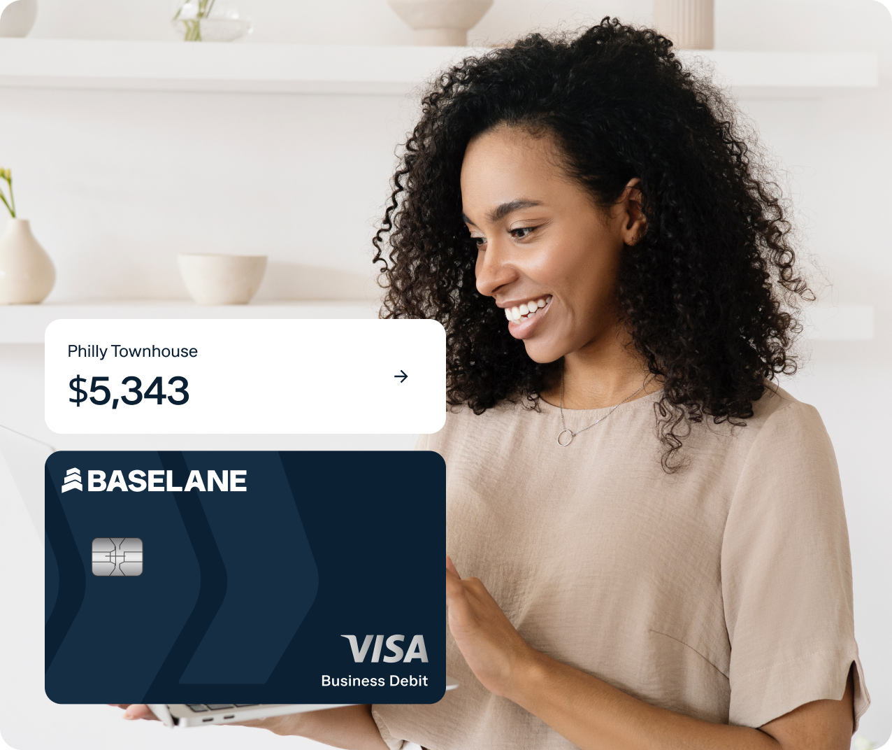 A smiling woman with curly hair is holding a tablet. A text overlay on the image shows "Philly Townhouse $5,343" with a right arrow, and a Baseline Visa Business Debit card is displayed in the foreground. She stands in a bright room with white shelves in the background.