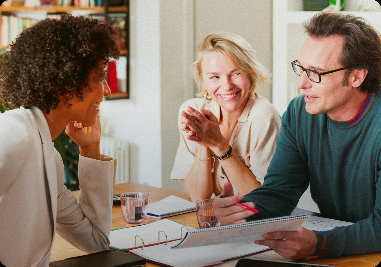 Three people are sitting at a table having a lively discussion. One person with curly hair is listening attentively, another person is smiling and clapping hands together, and the third person is holding a red pen and gesturing while talking. Documents are on the table.