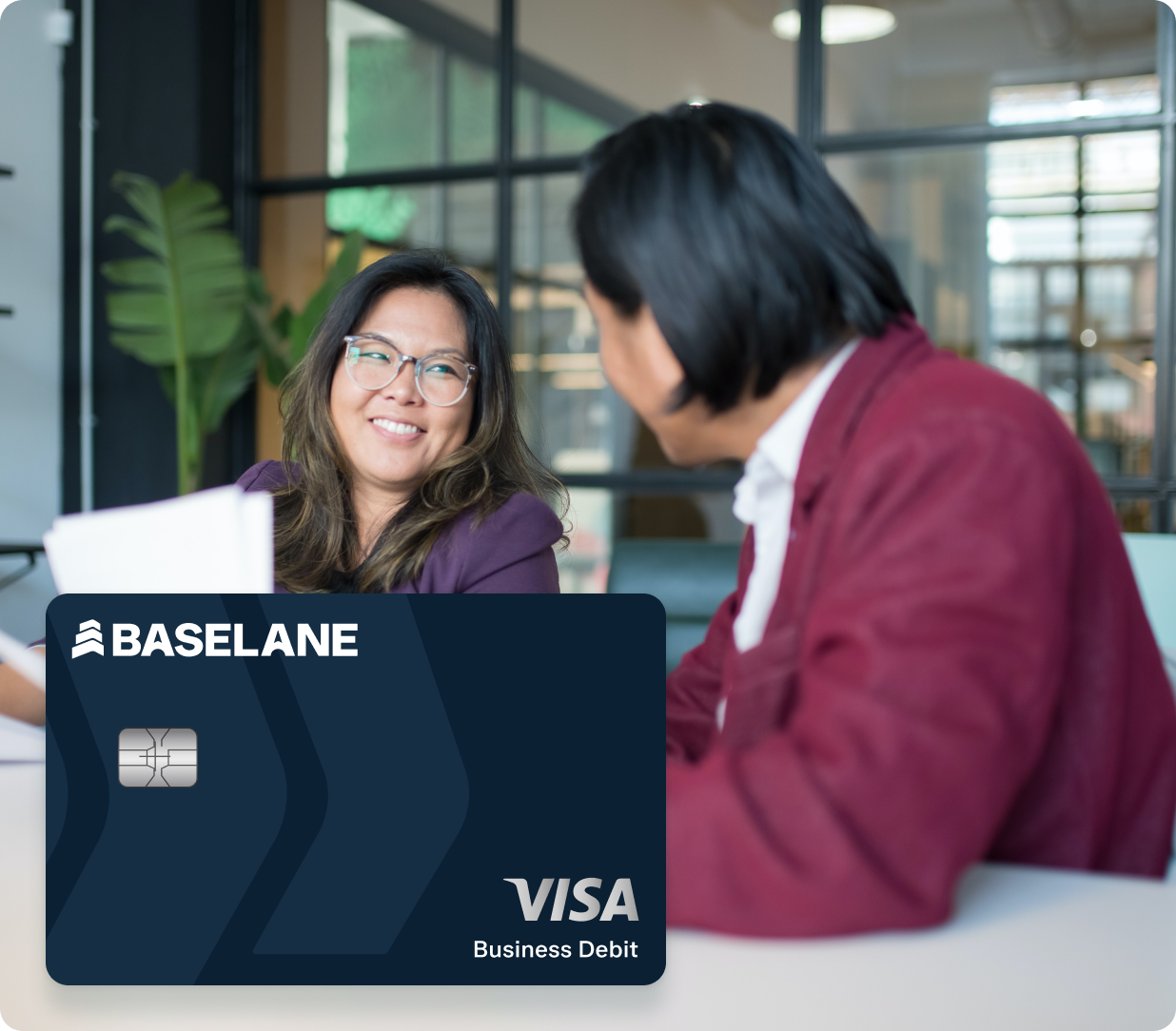 Two individuals are seated at a table having a discussion in a modern, glass-walled office space. One person, wearing glasses, is smiling. Overlaid in the foreground is a Baseline Visa Business Debit card.