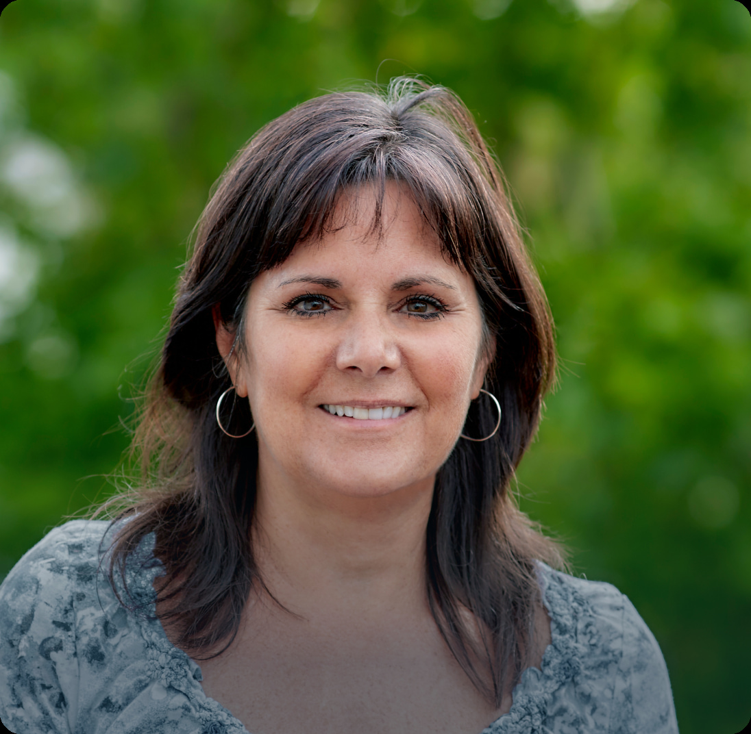 A woman with shoulder-length dark brown hair smiles warmly at the camera. She is wearing a patterned grey top and hoop earrings. The background is filled with lush green foliage, suggesting an outdoor setting.
