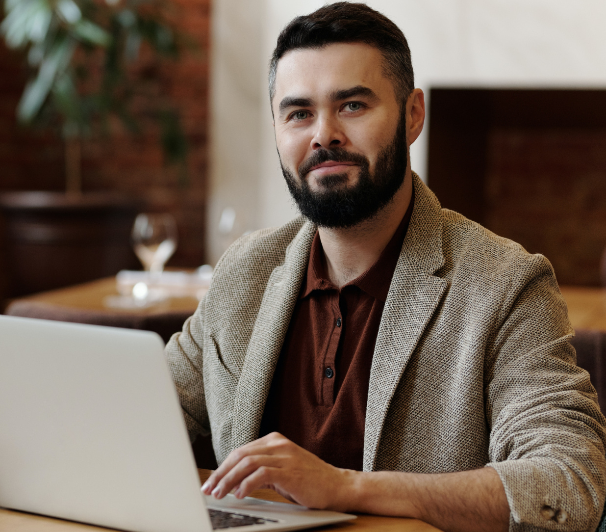 A man with a beard and short hair is sitting at a wooden table, using a laptop. He is wearing a brown polo shirt and a beige blazer. The background includes a plant and blurred dining items, suggesting a comfortable indoor environment.