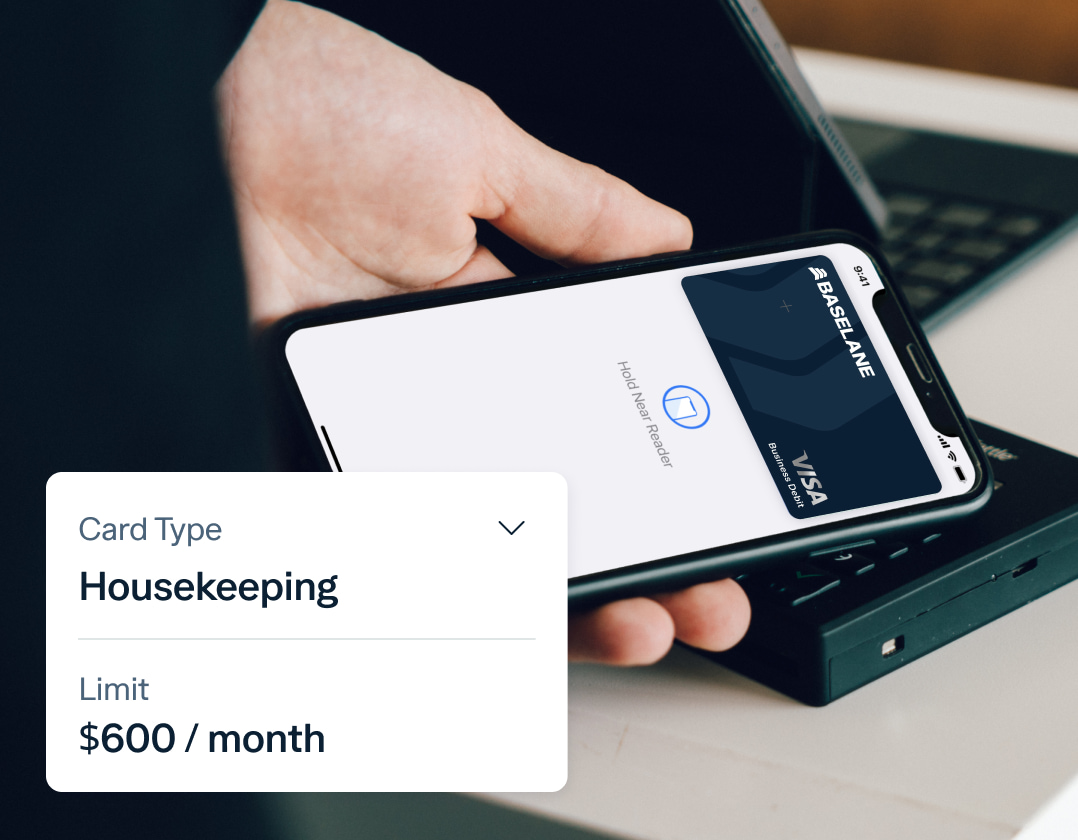 A person's hand holds a smartphone with a banking app open, displaying a virtual Visa card from Baseline for housekeeping. The card type is shown as Housekeeping with a limit of $600 per month. A card reader is visible in the background.