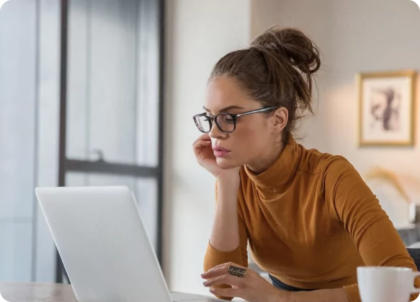 A woman with dark hair in a messy bun, wearing glasses and a brown turtleneck, is intently looking at a laptop screen. She is resting her chin on her hand, sitting at a desk with a cup beside her. The background shows a window and framed artwork on the wall.