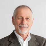 A man with short gray hair and a close-cropped beard is smiling softly. He is wearing a white collared shirt and a brown tweed blazer. The background is plain and light-colored.