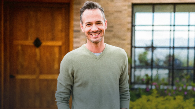 Man smiling outside a house with a wooden door and large window in the background. He is wearing a light green sweater and has short, slightly graying hair. The background includes a garden with flowers.