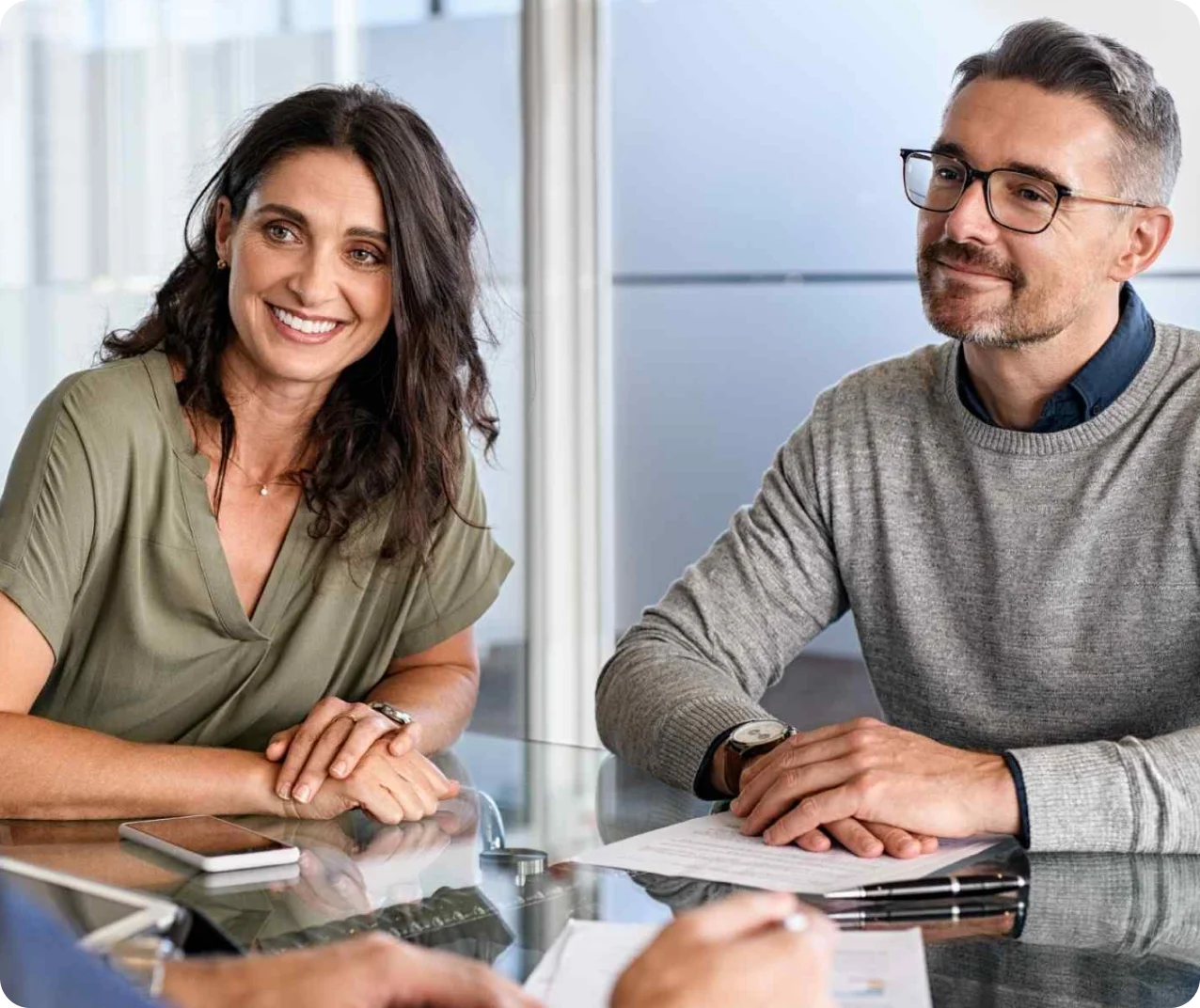 A woman and a man are seated at a glass table, engaged in a discussion. The woman has long brown hair and is smiling, wearing a green blouse. The man has short gray hair, glasses, and is wearing a gray sweater. Papers and a smartphone are on the table.