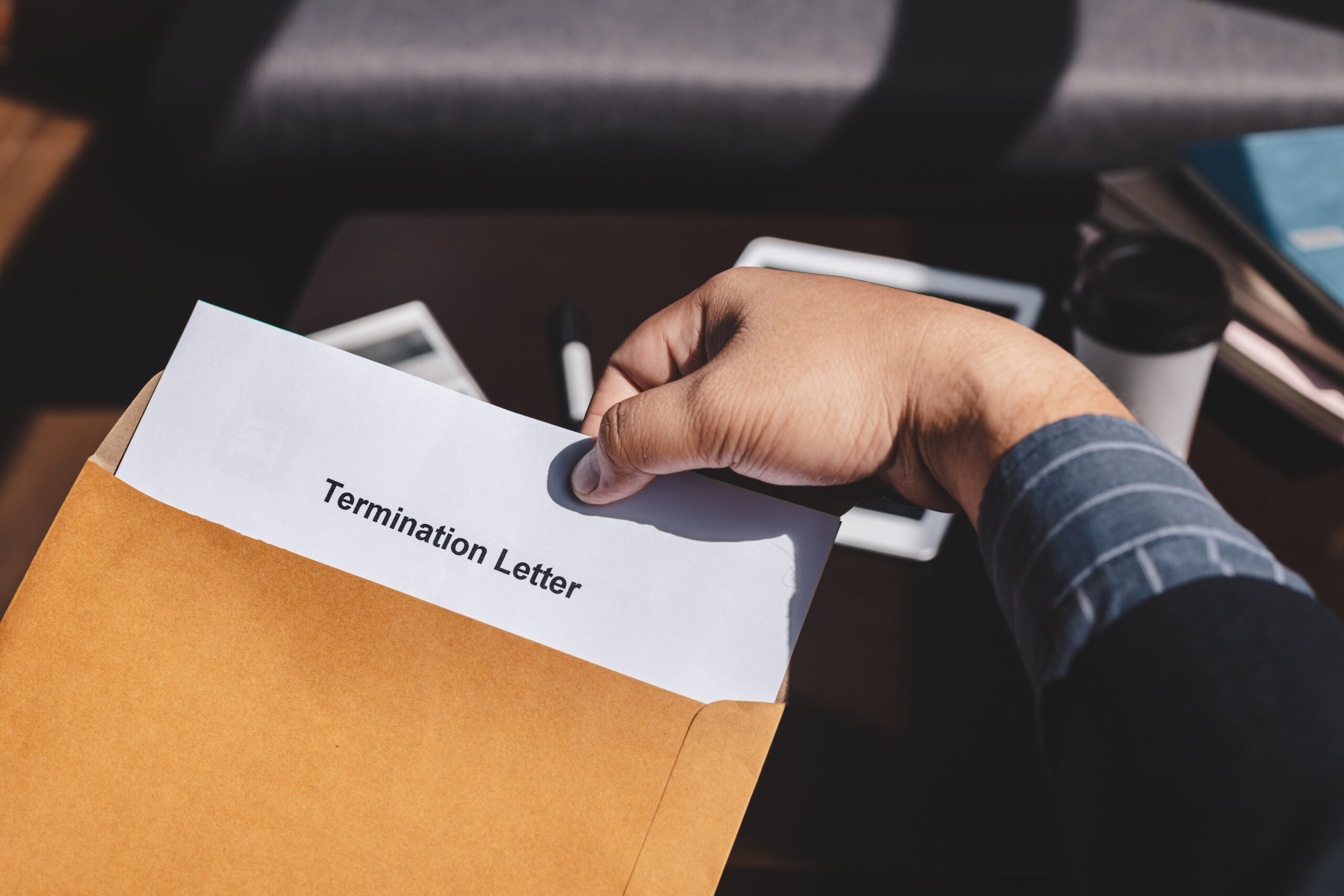 A person holds a brown envelope with a termination letter partially visible inside. The scene includes a desk with a tablet, a pen, and a coffee cup in the background.