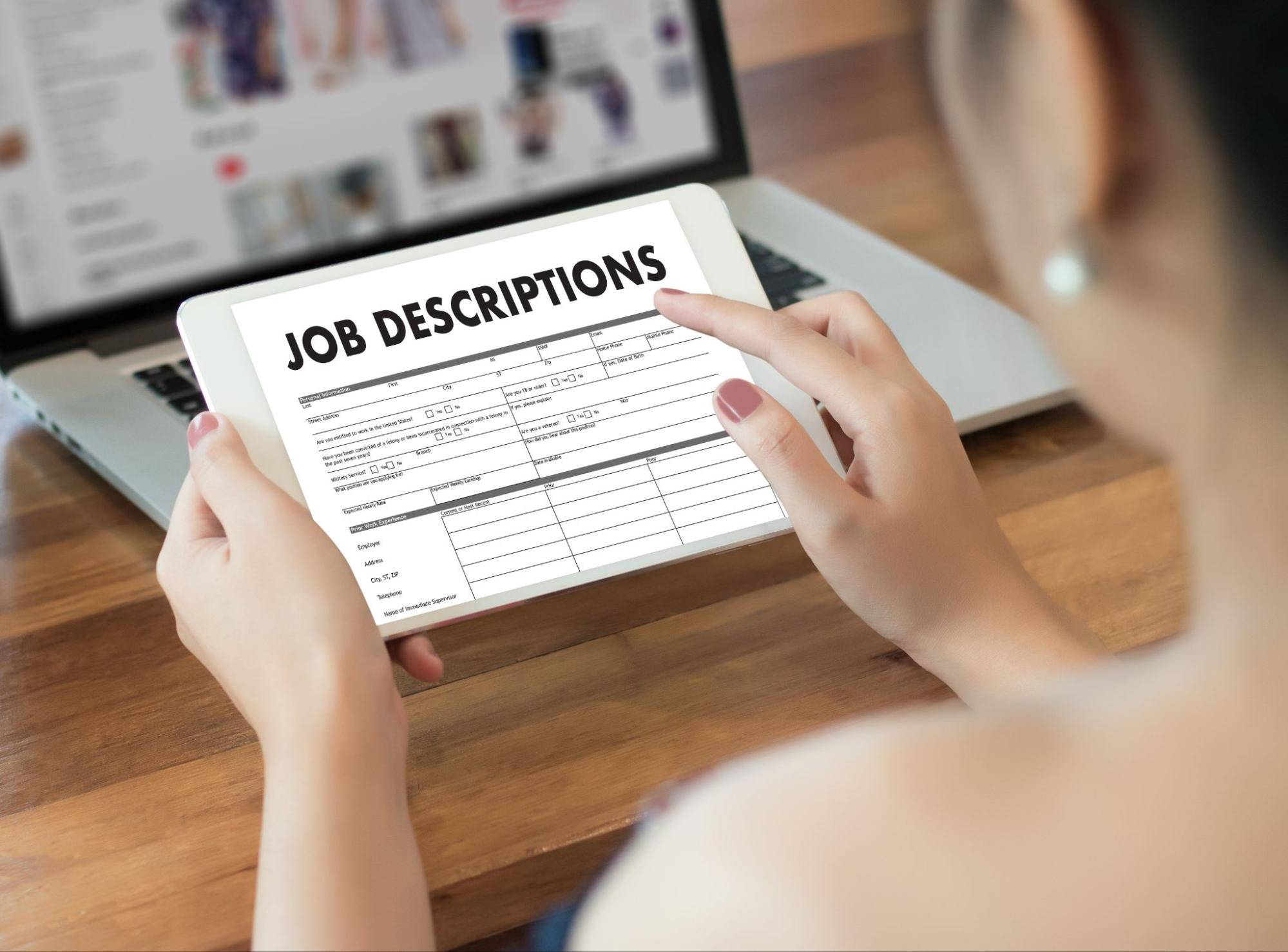 A person holds a tablet displaying a job description form. In the background, there's a laptop on a wooden table showing an online store. The scene suggests multitasking or job searching while browsing online.