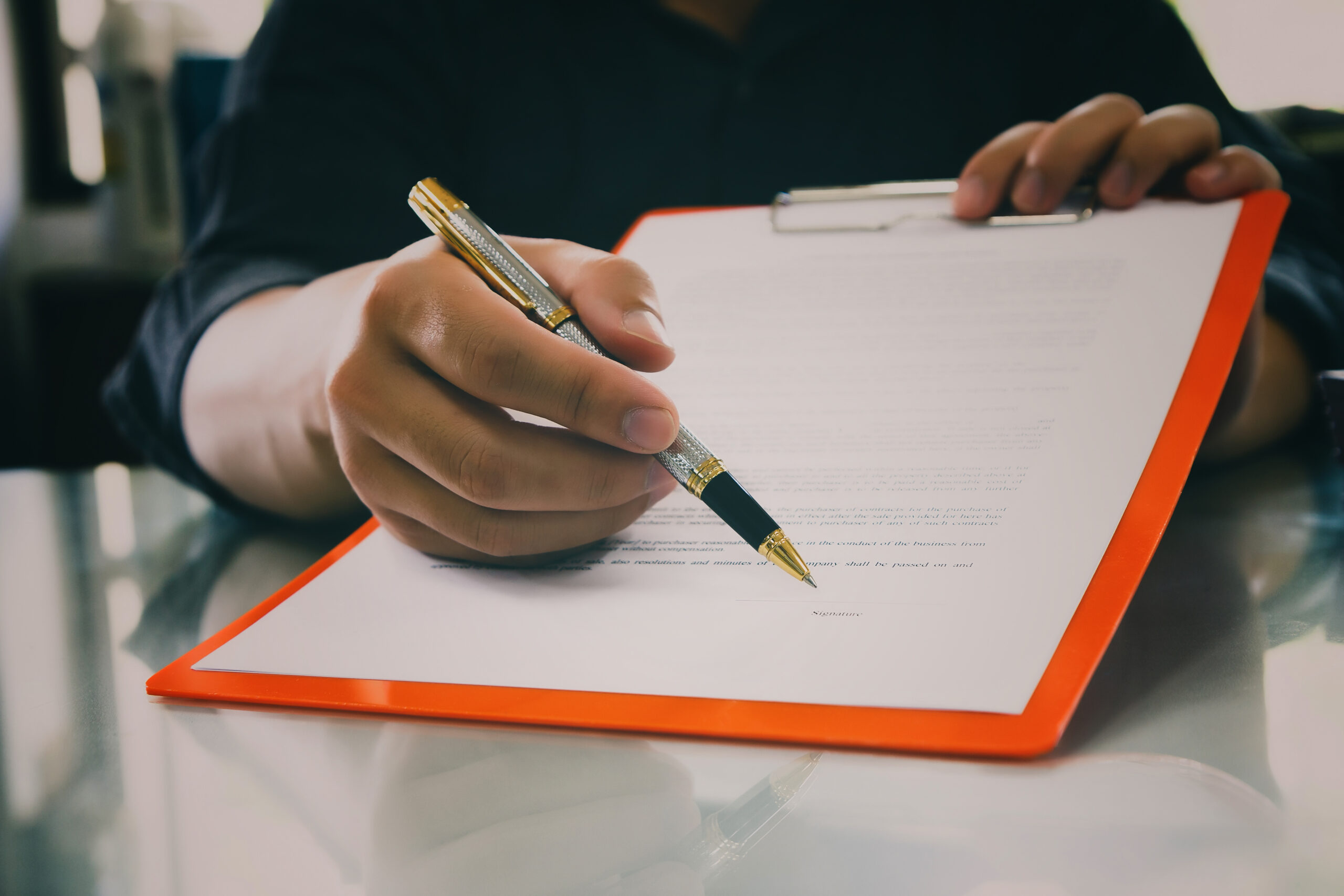 A person holding a pen is poised to write on a document clipped to a red clipboard. The individual is wearing a black shirt, and the scene is set on a reflective surface.