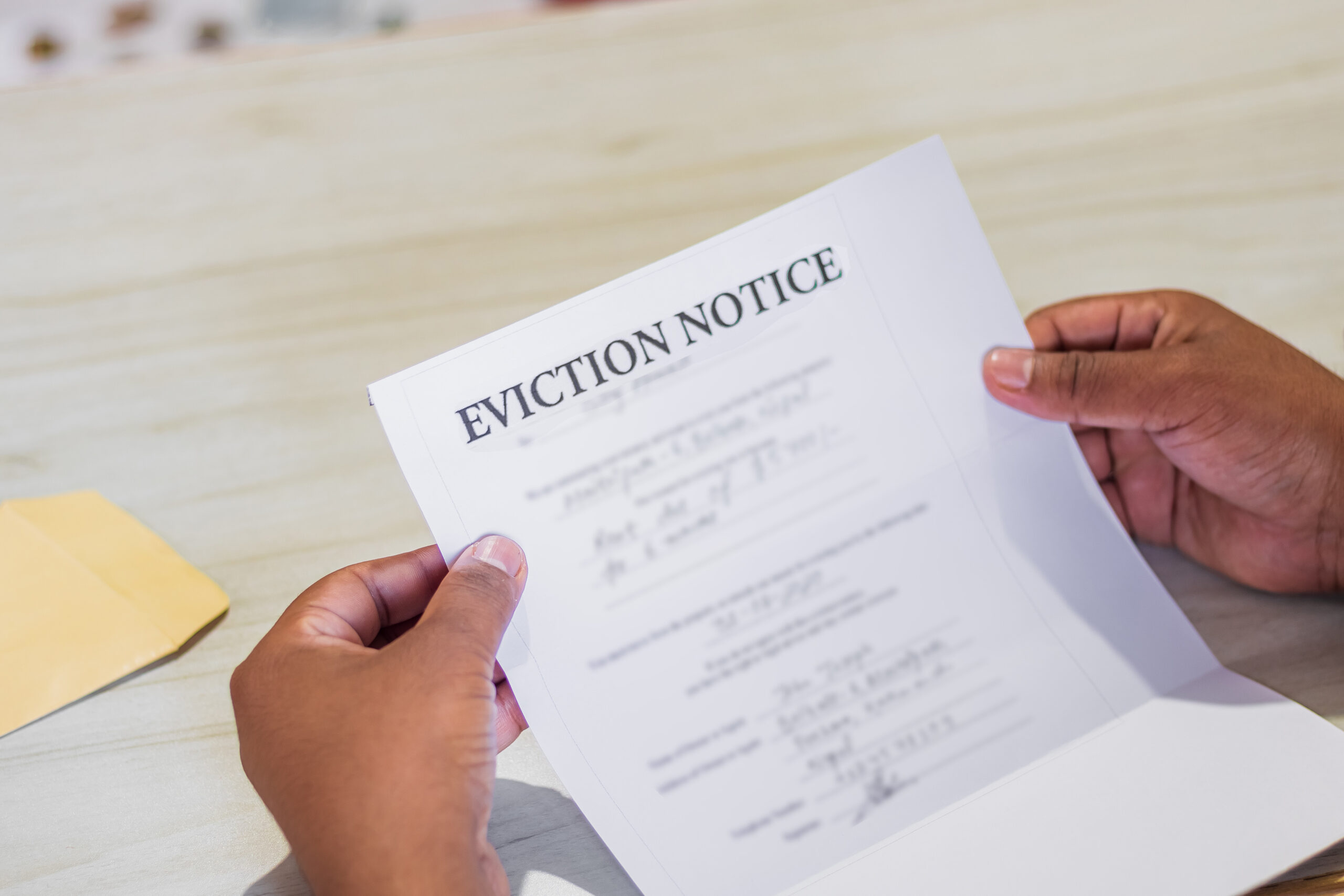 A person holding an eviction notice with both hands over a light-colored table. A yellow envelope is partially visible on the table in the background. The document is out of focus except for the bold heading that reads "EVICTION NOTICE.
