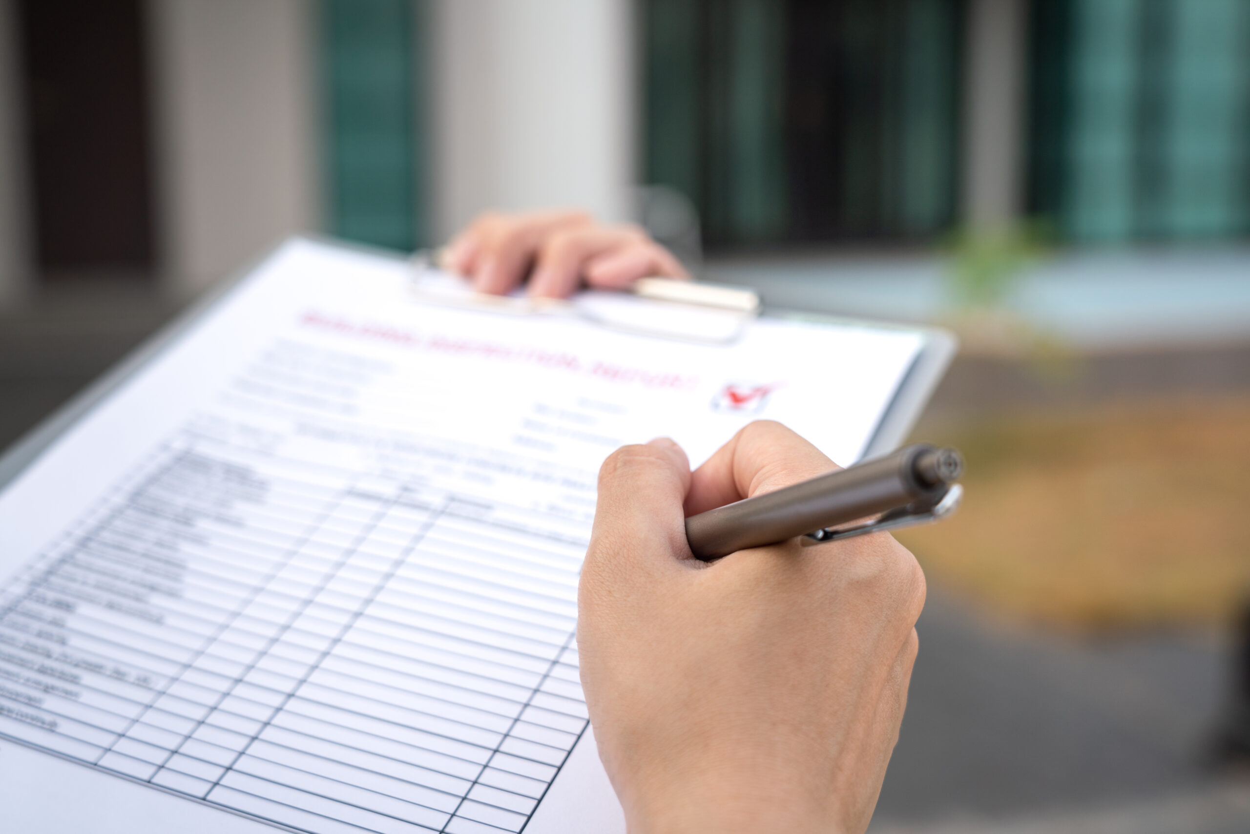 A person is holding a clipboard and writing with a pen. The clipboard contains a printed form, possibly for inspection or survey purposes. Blurry background features a building with green-tinted windows.