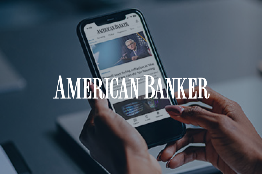 A person holds a smartphone displaying the "American Banker" website, which features a news article with an image of an older man wearing glasses. The background includes a blurred laptop.