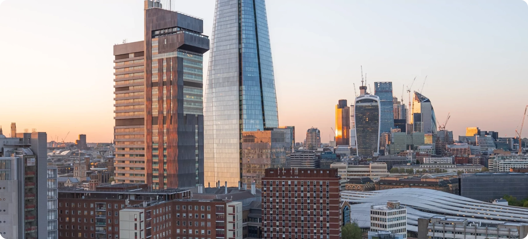 London skyline with modern skyscrapers at sunset, including the Shard, surrounded by other buildings. The sky is clear with a warm glow, creating a contrast between the glass structures and the older architecture.