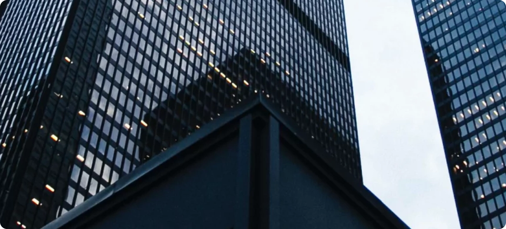 View of a tall, glass-covered skyscraper from a low angle. The building reflects nearby structures and the cloudy sky, creating a geometric pattern. The structure's sharp angles dominate the foreground, emphasizing its modern architectural design.