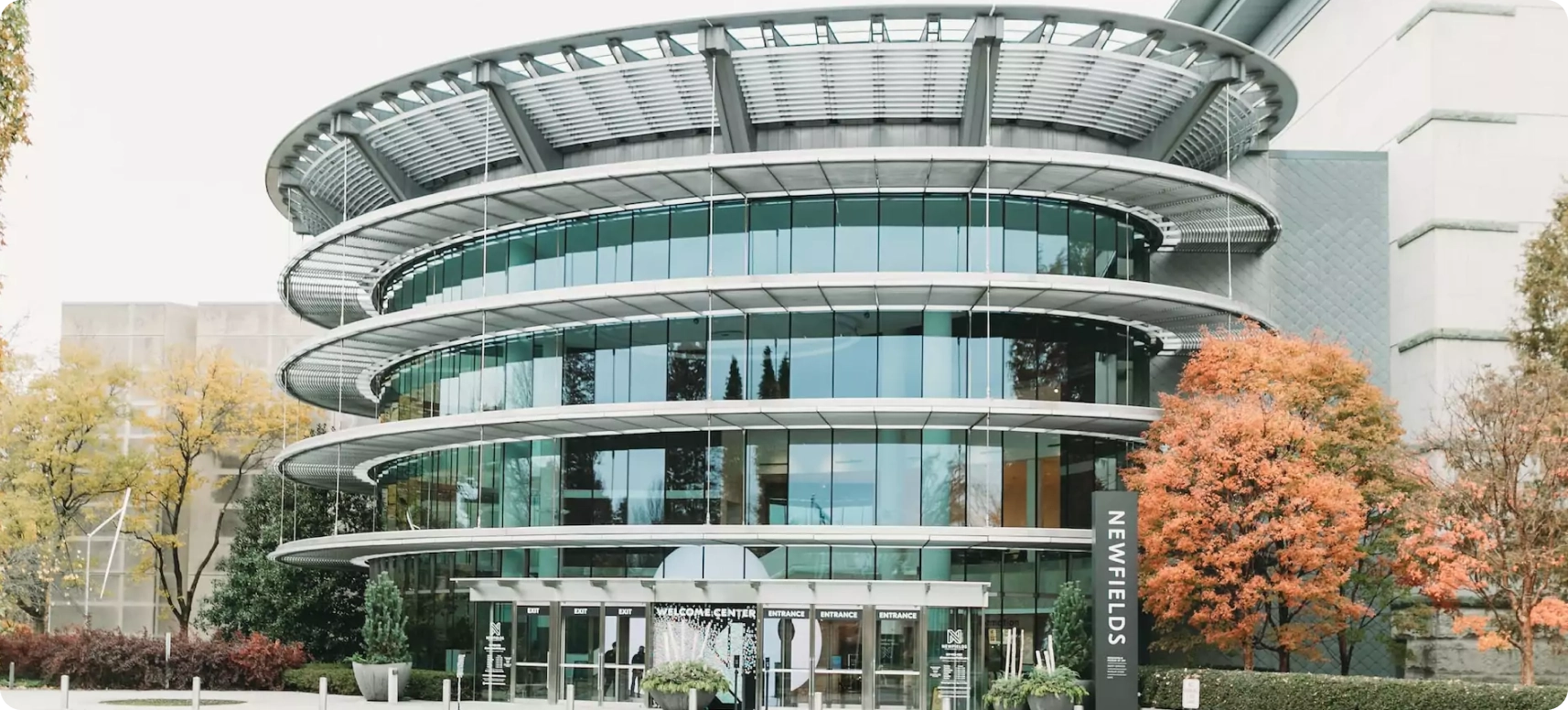 A modern glass and steel circular building with multiple tiers and a curved roof, surrounded by autumnal trees. The entrance features glass doors and decorations with the sign "Newfields" visible on the right.