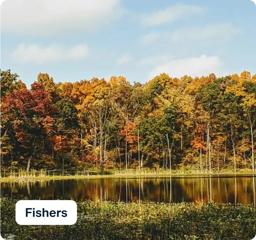 A serene lake reflects a dense forest with vibrant autumn foliage in shades of orange, red, and yellow under a partly cloudy sky. A label in the bottom corner reads "Fishers.