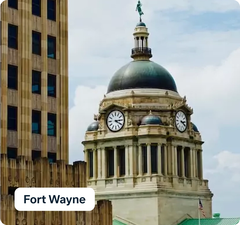 A historic clock tower with a dome and a statue on top is surrounded by modern buildings. A sign in the foreground reads "Fort Wayne." The sky is partly cloudy.