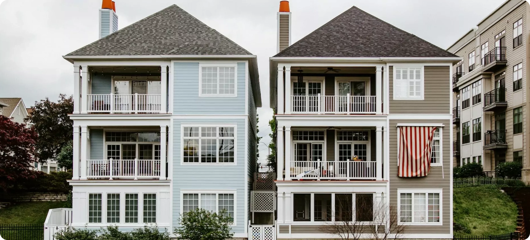 Two adjacent three-story houses with balconies and chimneys. The house on the left is light blue, while the one on the right is beige with a red and white striped awning. Both have white railings and windows, set against a cloudy sky.