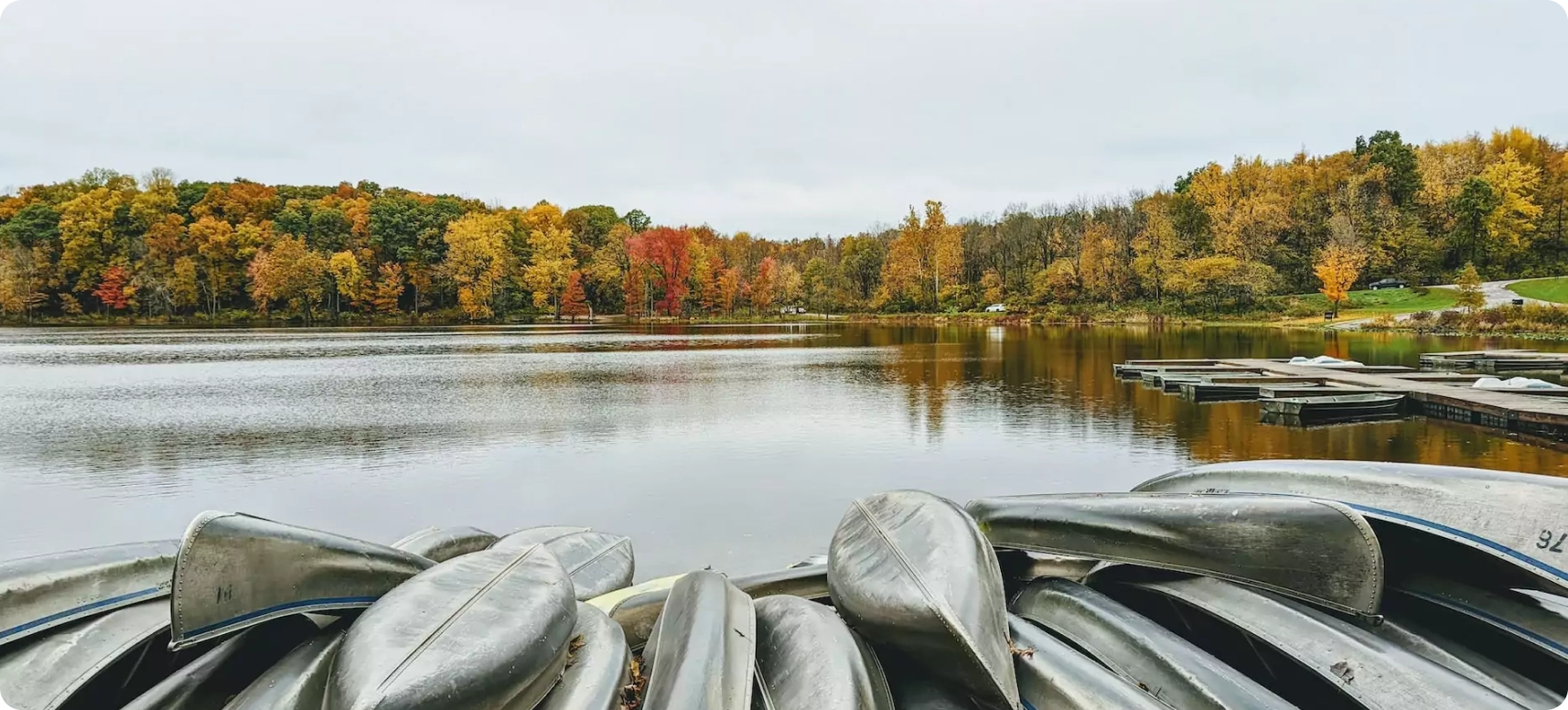 A serene lake lined with colorful autumn trees, reflecting vibrant red, orange, and yellow hues in the calm water. Canoes are stacked in the foreground, and a few docks extend into the lake. The sky is overcast.