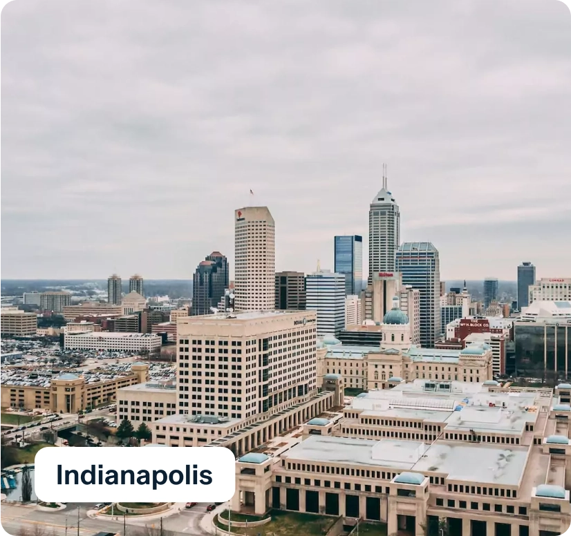 Aerial view of downtown Indianapolis, featuring a skyline with tall buildings under a cloudy sky. A mix of modern and classic architecture is visible, including glass skyscrapers and historic structures. A text label reads "Indianapolis.