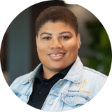 A person with short hair is smiling, wearing a denim jacket over a black shirt. The background is softly blurred, featuring some greenery and gentle lighting.