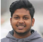 A person with short dark hair and a slight smile is wearing a gray hoodie. The background is blurred, suggesting an outdoor setting.
