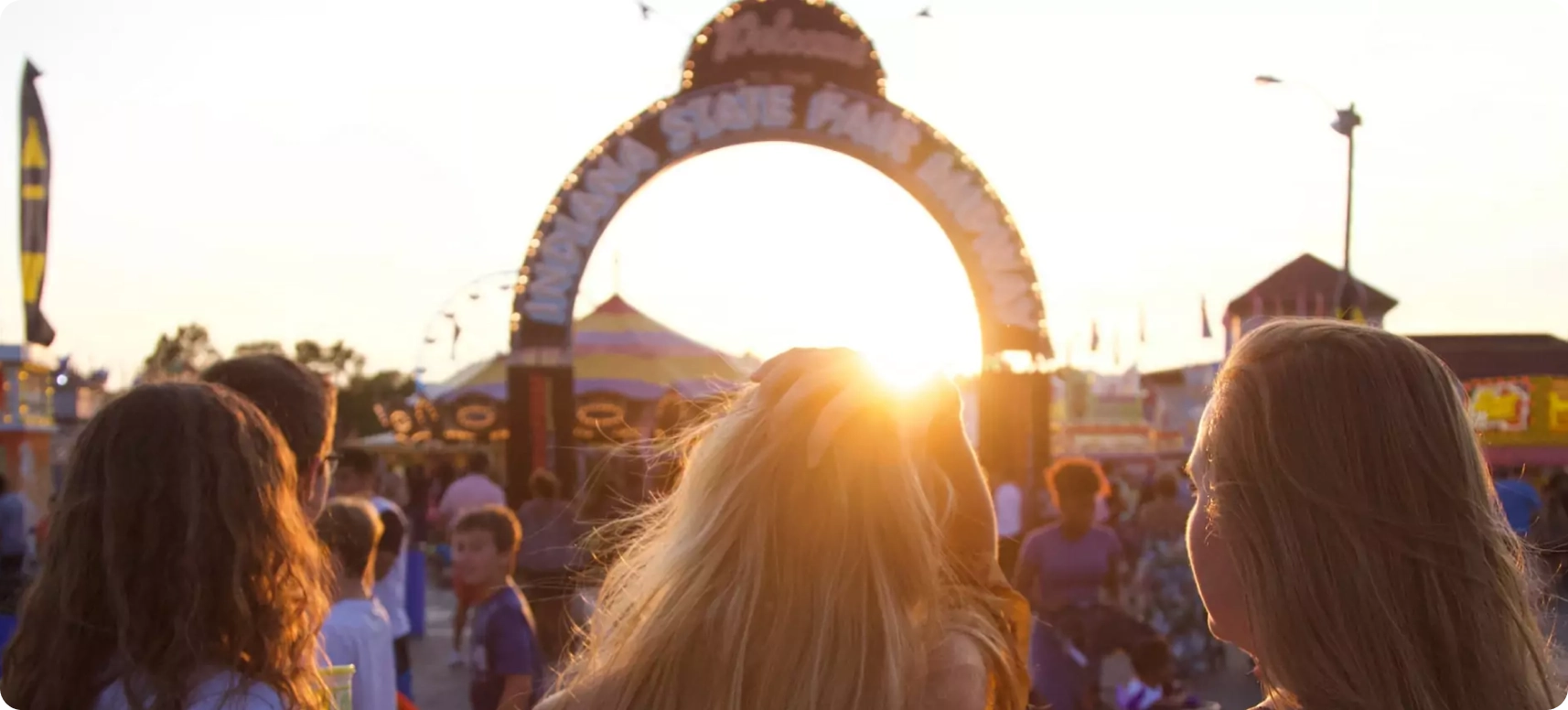 Three people are walking towards a sunlit entrance arch at a lively festival. The background shows a crowd of people, tents, and flags. The sky is bright as the sun sets behind the arch.