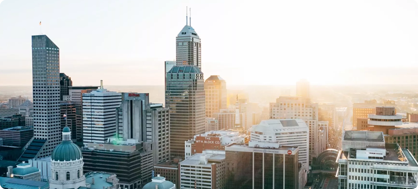 Aerial view of a city skyline at sunrise, featuring a collection of modern skyscrapers and high-rise buildings. Sunlight glows in the background, casting warm hues over the cityscape.