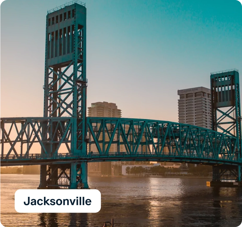 A blue steel drawbridge over a calm river at sunset, with tall city buildings in the background. The sky is a gradient of orange to blue. The word "Jacksonville" is displayed on the bottom left.