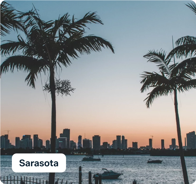 Silhouette of a city skyline with tall buildings at dusk, framed by two palm trees. The sky displays soft orange and blue hues, and a calm body of water is visible in the foreground with boats. A label reads "Sarasota.