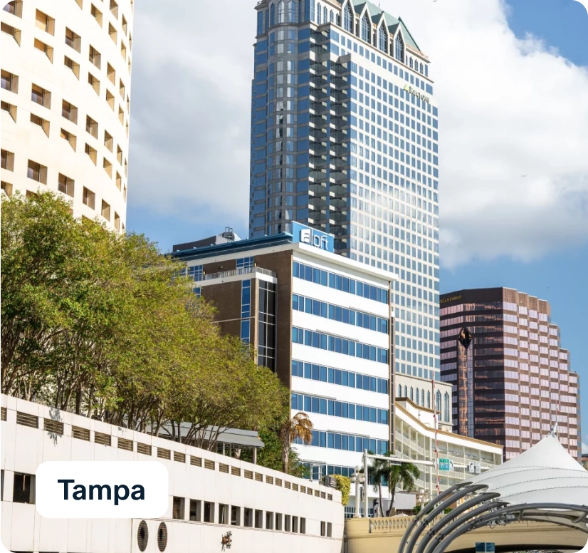View of downtown Tampa, highlighting several modern skyscrapers under a partly cloudy sky. The foreground features a row of trees and a portion of a curved structure on the right. A white sign with "Tampa" is visible.