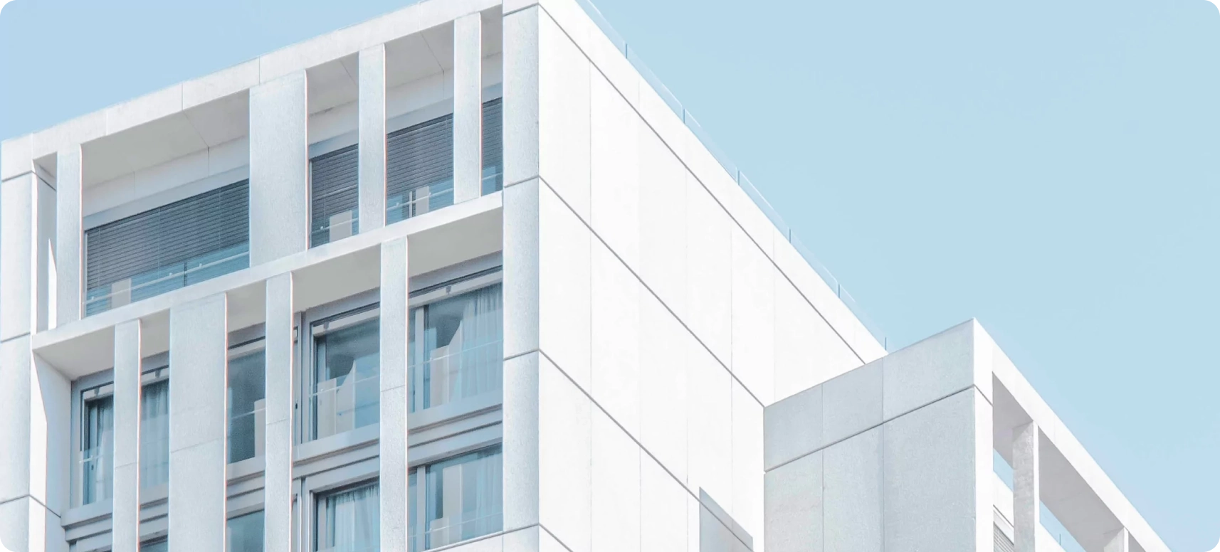 A minimalist, modern white building with large windows and clean lines stands against a clear blue sky. The architecture features rectangular shapes and a smooth facade.