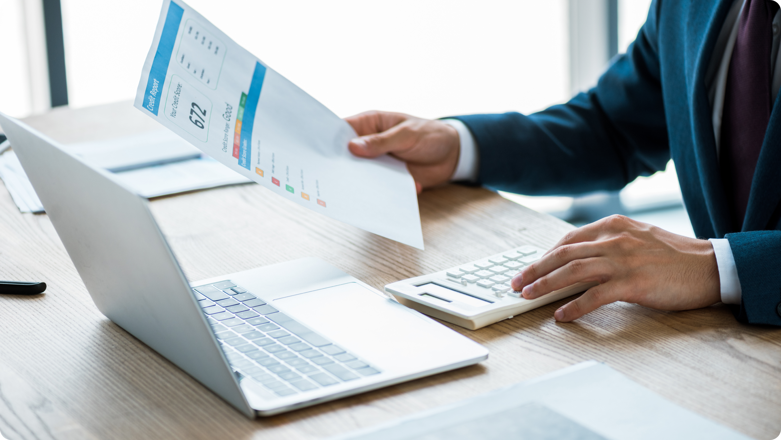 A landlord in a suit sits at a desk, working with a laptop and a calculator. They hold a printed credit report displaying a tenant's credit check details.