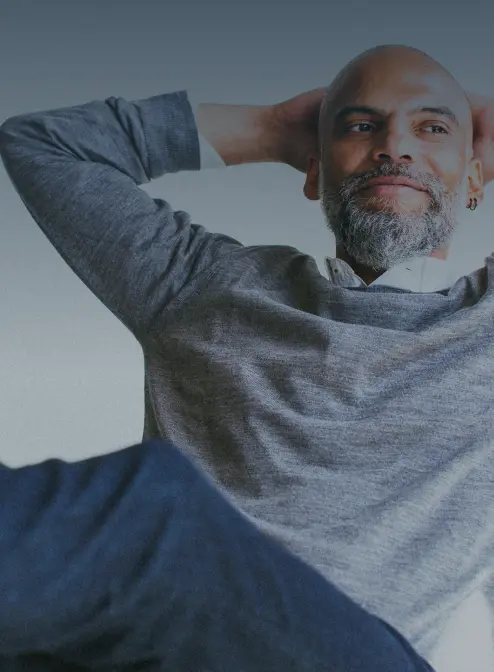 A relaxed person with a bald head and beard leans back with hands behind their head, wearing a gray sweater and blue pants. They appear content and are looking slightly upwards.