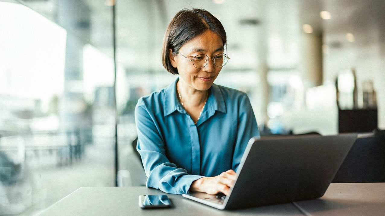 A woman in glasses and a teal blouse sits at a table working on a laptop. Her smartphone is beside her. The background is a bright, modern indoor space with blurred details.