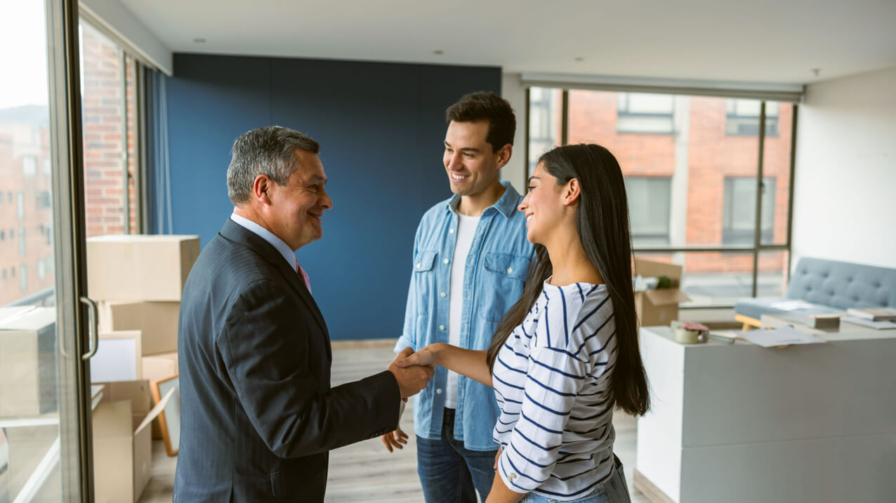 A real estate agent in a suit shakes hands with a smiling couple in casual clothes inside a sunlit room. Moving boxes are visible in the background near a large window with a city view.