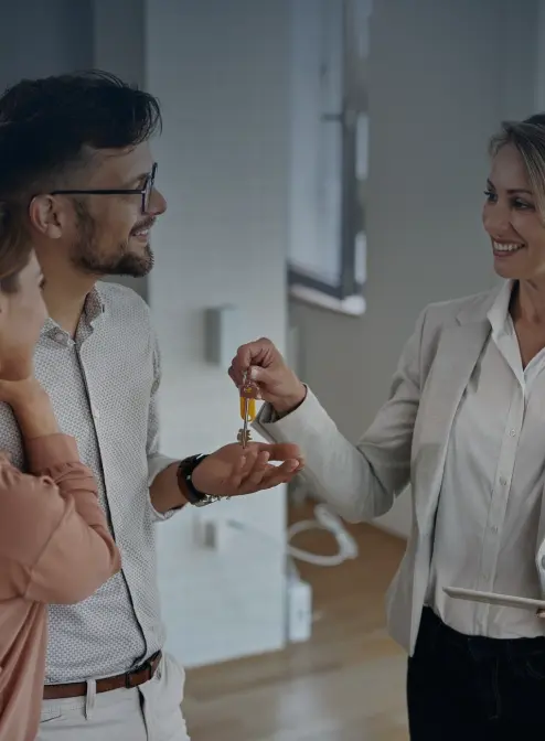 A real estate agent hands a set of keys to a smiling couple inside an empty room. The couple appears happy and excited, suggesting a new home purchase or rental agreement. The room is minimally furnished, indicating a recent move.