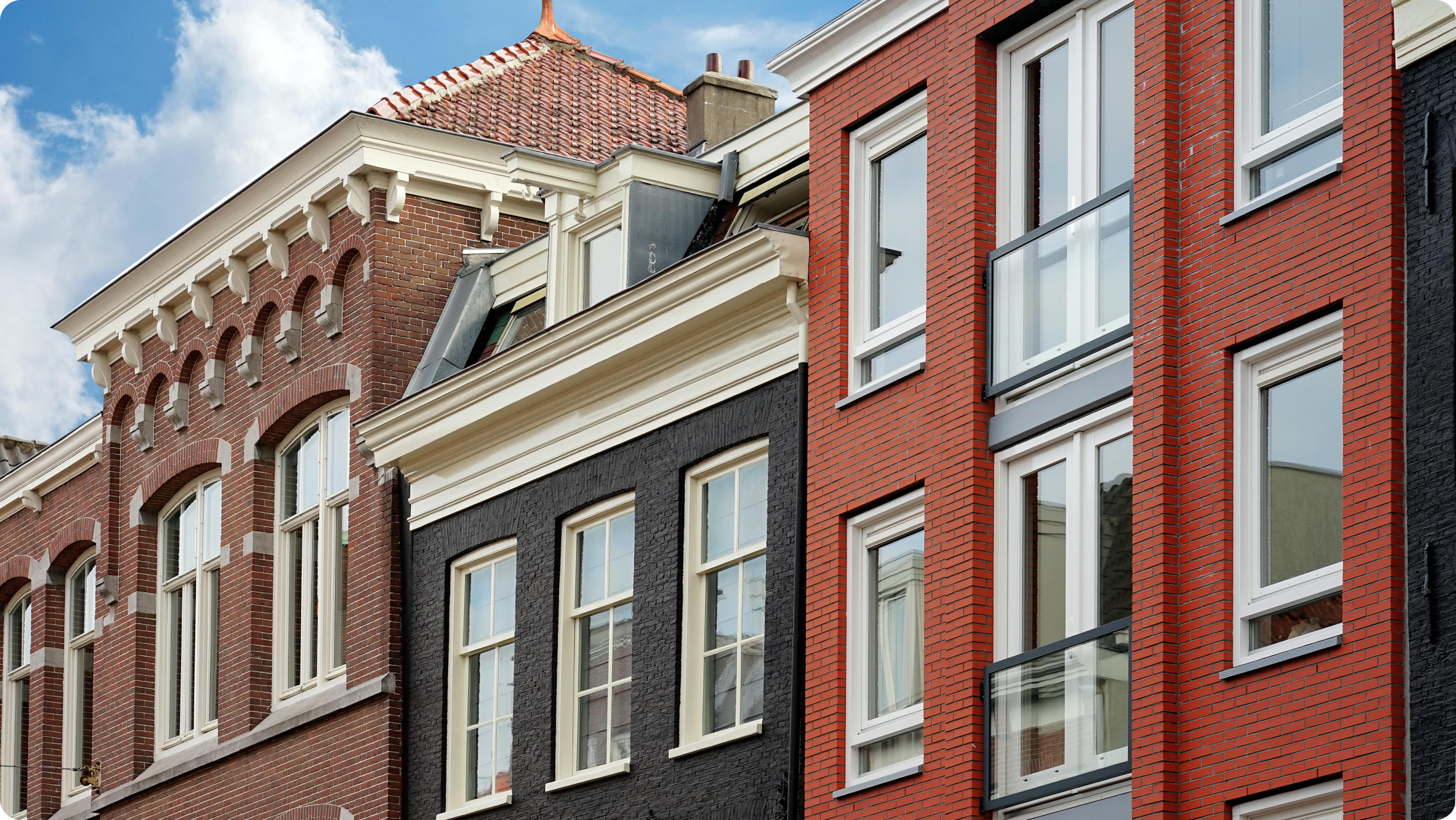 A row of European-style buildings with tall windows and intricate brickwork. One building is black with white trim and arched windows, while the adjacent one is red with rectangular white-trimmed windows, set against a cloudy blue sky.